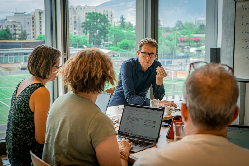 Daniel Schwenger spricht mit drei Teilnehmenden an einem Tisch mit Laptops